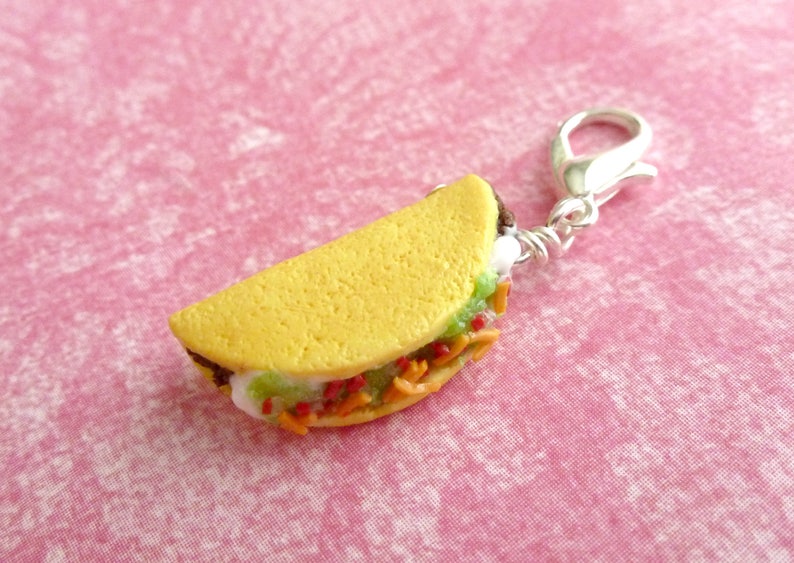May include: A small, yellow, clay taco charm with a silver clasp. The taco is filled with green lettuce, red tomatoes, and orange cheese.