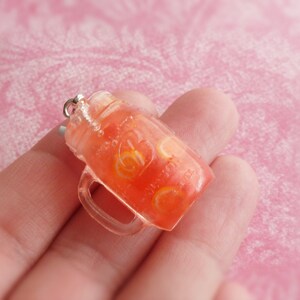 May include: A clear glass jar charm filled with a red and orange liquid, resembling a fruit-flavoured drink. The jar has a silver lid and a clear handle.