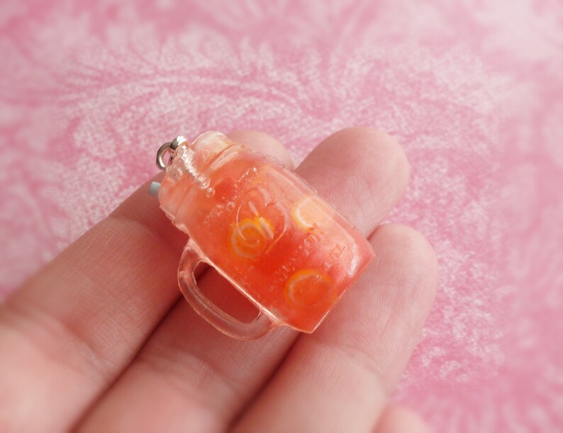 May include: A clear glass jar charm filled with a red and orange liquid, resembling a fruit-flavoured drink. The jar has a silver lid and a clear handle.