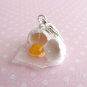 May include: A silver charm featuring a white egg with a broken shell and a yellow yolk inside. The charm is attached to a silver chain.