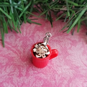 May include: A miniature red mug charm filled with hot chocolate, topped with marshmallows and chocolate drizzle. The charm has a silver-colored loop for attaching to a bracelet or necklace. The charm is on a pink background.