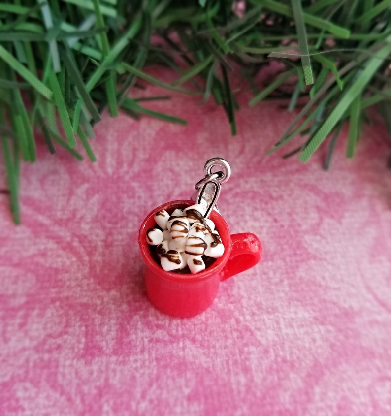 May include: A miniature red mug charm filled with hot chocolate, topped with marshmallows and chocolate drizzle. The charm has a silver-colored loop for attaching to a bracelet or necklace. The charm is on a pink background.