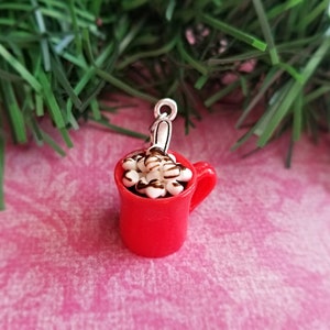 May include: A miniature red mug charm filled with white marshmallows drizzled with chocolate. The charm has a silver-colored loop for attaching to a necklace or bracelet. The mug has a handle and is set against a pink background.