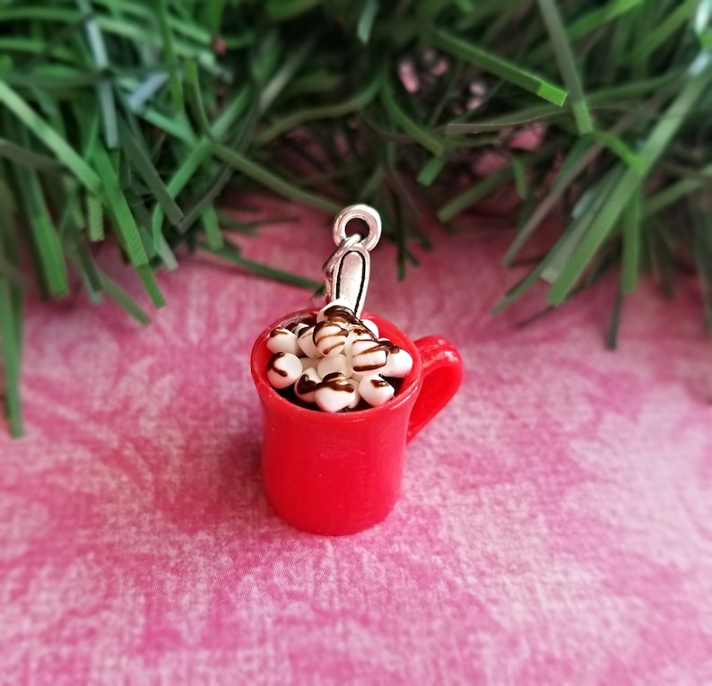 May include: A miniature red mug charm filled with white marshmallows drizzled with chocolate. The charm has a silver-colored loop for attaching to a necklace or bracelet. The mug has a handle and is set against a pink background.