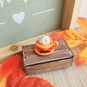 May include: A miniature orange ceramic coffee cup with whipped cream and a sprinkle of cinnamon on top, sitting on a small wooden crate. The cup is on a saucer with a cinnamon stick.