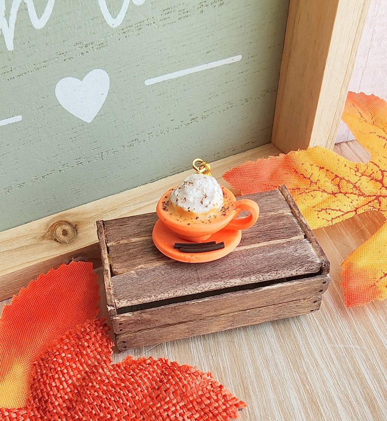 May include: A miniature orange ceramic coffee cup with whipped cream and a sprinkle of cinnamon on top, sitting on a small wooden crate. The cup is on a saucer with a cinnamon stick.