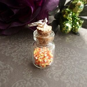 May include: A small, clear glass bottle filled with candy corn. The bottle has a cork stopper and a silver-colored charm attached to the top. The candy corn is yellow, orange, and white. The background is a blurred floral design.