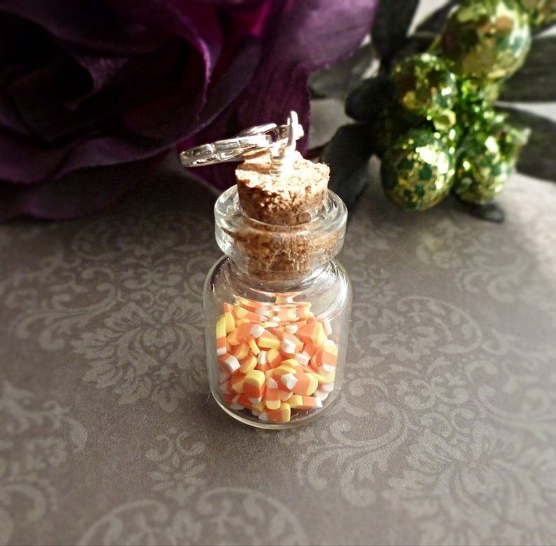 May include: A small, clear glass bottle filled with candy corn. The bottle has a cork stopper and a silver-colored charm attached to the top. The candy corn is yellow, orange, and white. The background is a blurred floral design.