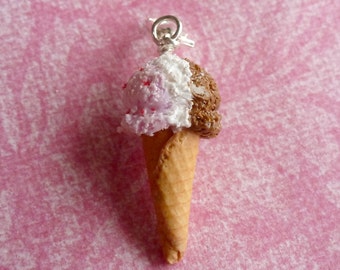 May include: A miniature ice cream cone charm with a silver-colored loop. The ice cream has a split design, with white and pink ice cream on one side and brown ice cream on the other. The cone is a light brown color.