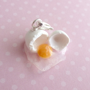 May include: A white ceramic egg charm with a silver loop, cracked open to reveal a yellow yolk. The egg is sitting on a white background.