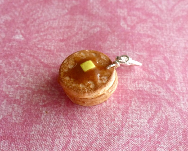 May include: A charm of a stack of two miniature pancakes with a yellow pat of butter on top. The charm is made of polymer clay and has a silver jump ring.