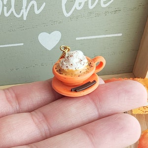 May include: A miniature orange ceramic coffee cup with whipped cream and cinnamon sprinkles, on a saucer with chocolate sticks, and a gold chain attached.
