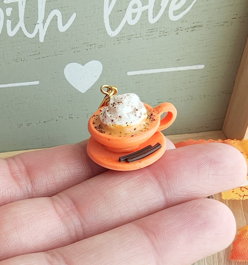 May include: A miniature orange ceramic coffee cup with whipped cream and cinnamon sprinkles, on a saucer with chocolate sticks, and a gold chain attached.