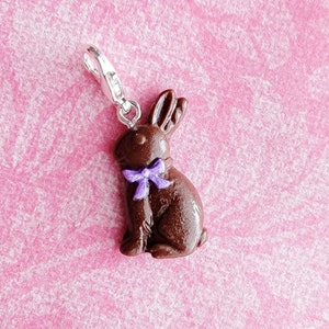 May include: A chocolate-colored bunny charm with a purple bow, attached to a silver-colored clasp. The bunny charm is a detailed, three-dimensional design. The background is a pink, textured surface.