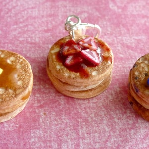 May include: Three miniature pancake charms with different toppings. The first pancake has a yellow pat of butter and brown syrup. The second pancake has red strawberry slices and red syrup. The third pancake has blue blueberries and brown syrup.