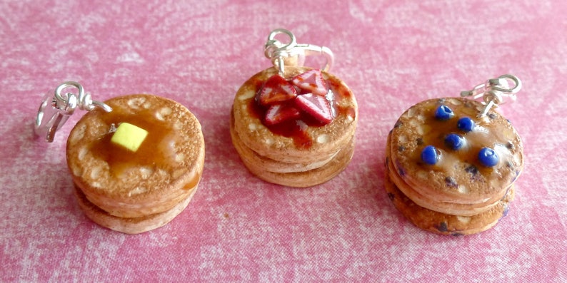 May include: Three miniature pancake charms with different toppings. The first pancake has a yellow pat of butter and brown syrup. The second pancake has red strawberry slices and red syrup. The third pancake has blue blueberries and brown syrup.