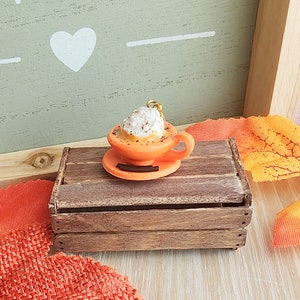 May include: A miniature orange ceramic mug with a white whipped cream topping and a sprinkle of cinnamon. The mug is sitting on a brown wooden crate.