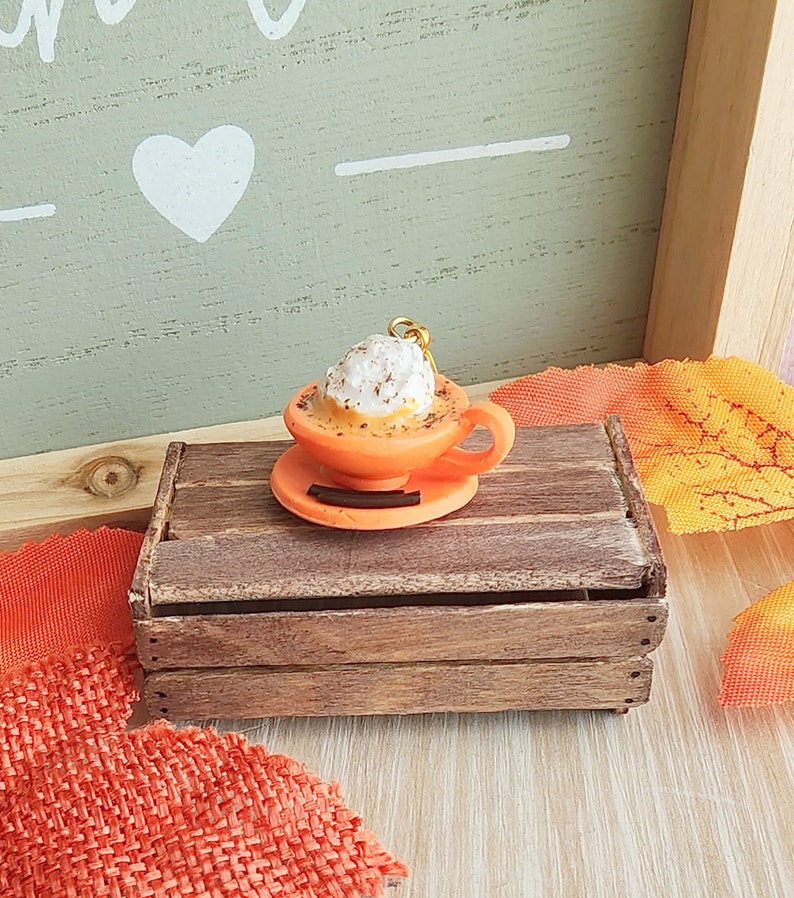 May include: A miniature orange ceramic mug with a white whipped cream topping and a sprinkle of cinnamon. The mug is sitting on a brown wooden crate.