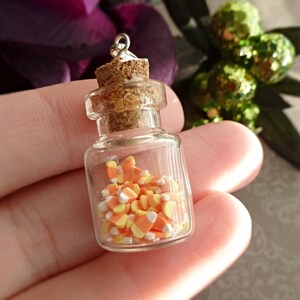 May include: A miniature glass bottle filled with candy corn. The bottle has a cork stopper and a silver loop at the top. The candy corn is orange, yellow, and white. The bottle is held in a person's hand.