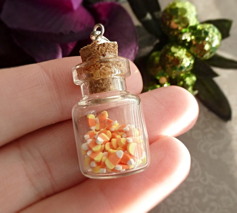 May include: A miniature glass bottle filled with candy corn. The bottle has a cork stopper and a silver loop at the top. The candy corn is orange, yellow, and white. The bottle is held in a person's hand.