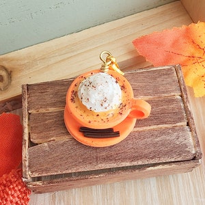 May include: A small orange and white ceramic charm of a coffee cup with whipped cream and sprinkles. The charm is hanging from a gold chain and is sitting in a wooden box.