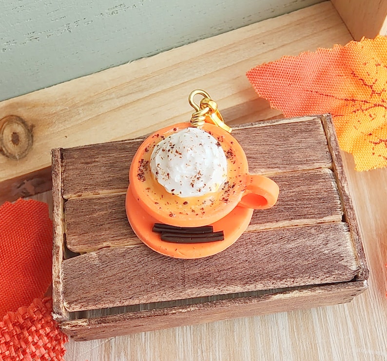 May include: A small orange and white ceramic charm of a coffee cup with whipped cream and sprinkles. The charm is hanging from a gold chain and is sitting in a wooden box.