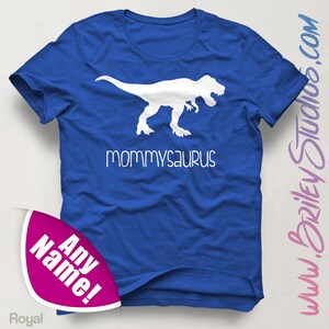 May include: A royal blue t-shirt with a white dinosaur silhouette and the text "mommysaurus" printed on the front. The shirt has a folded sleeve and is on a white background.