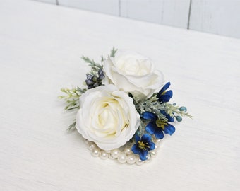 Puede incluir: Un ramillete floral con dos rosas blancas, flores azules y follaje. El arreglo está adornado con pequeñas perlas redondas. Ideal para bodas, bailes de graduación u ocasiones especiales. Un hermoso accesorio.