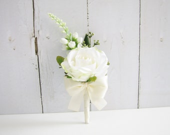 Puede incluir: Un boutonniere de rosa blanca con un lazo de satén blanco y follaje verde.