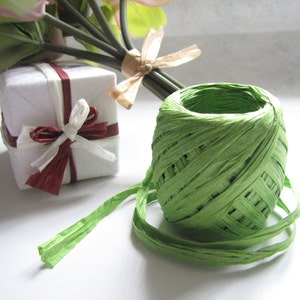 May include: A spool of green paper twine with a white gift box and a bouquet of pink and white flowers in the background.