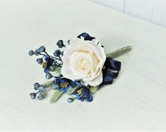 Puede incluir: Un prendido de rosa blanca con detalles azul marino. El arreglo floral incluye bayas azules, flores azules y amarillas y una cinta azul marino. El tallo está envuelto en cinta floral verde.