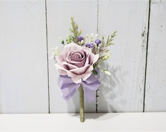 Op de afbeelding: Een enkele, lichtroze roos met een lavendelkleurige strik en groen. De roos is omringd door kleine witte en paarse bloemen.