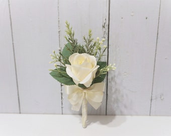 Puede incluir: Una sola rosa blanca con hojas verdes y pequeñas flores blancas, atadas con una cinta de satén blanca. El ramo está dispuesto sobre un fondo blanco.