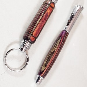 May include: A set of two wooden pens with a silver finish. The pen on the left is a keychain with a silver ring. The pen on the right is a full-sized pen. Both pens have a wood grain pattern with red, purple, and green colours.