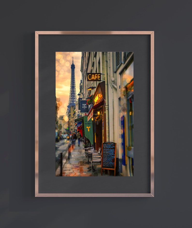 May include: A framed photograph of a Parisian street scene with a cafe and the Eiffel Tower in the background. The cafe has a sign that reads "CAFE" in yellow letters. There are tables and chairs set up outside the cafe.