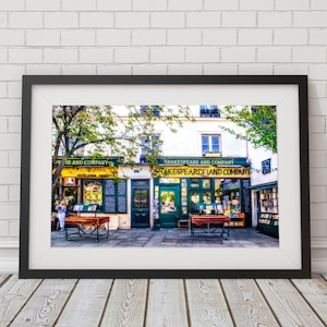 May include: A black framed photograph of two bookstores in Paris, France. The storefronts are green and yellow with the text "Shakespeare and Company" above the entrance. The stores are lined with bookshelves filled with books.