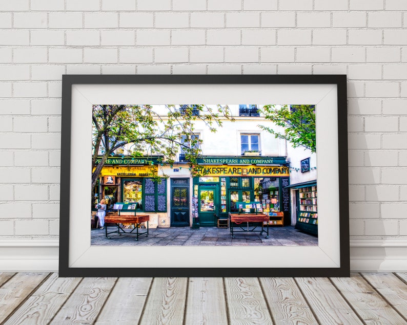 May include: A black framed photograph of two bookstores in Paris, France. The storefronts are green and yellow with the text "Shakespeare and Company" above the entrance. The stores are lined with bookshelves filled with books.