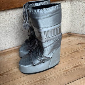 May include: A pair of silver Moon Boots with the brand name printed on the side. The boots have a shiny, metallic finish, thick soles, and laces. The boots are designed for cold weather and snow.