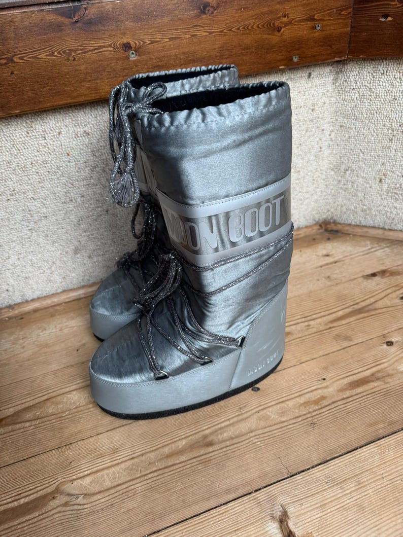 May include: A pair of silver Moon Boots with the brand name printed on the side. The boots have a shiny, metallic finish, thick soles, and laces. The boots are designed for cold weather and snow.