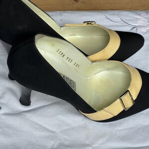 May include: A pair of black heels with pointed toes and a light tan strap across the top, secured with a small buckle. The heels have a slender, black heel. The shoes are on a white surface.