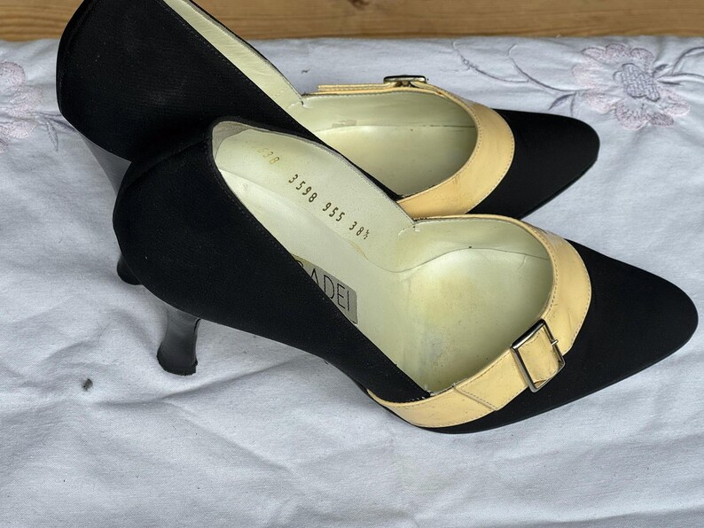 May include: A pair of black heels with pointed toes and a light tan strap across the top, secured with a small buckle. The heels have a slender, black heel. The shoes are on a white surface.