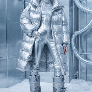 May include: A futuristic silver outfit, including a long puffer coat, a jumpsuit, and snow boots. The ensemble is paired with silver sunglasses. The outfit is designed for cold weather and has a shiny, metallic finish.