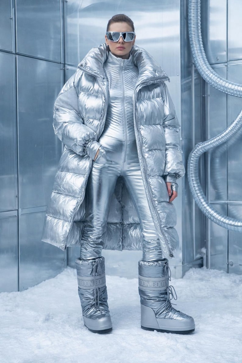 May include: A futuristic silver outfit, including a long puffer coat, a jumpsuit, and snow boots. The ensemble is paired with silver sunglasses. The outfit is designed for cold weather and has a shiny, metallic finish.