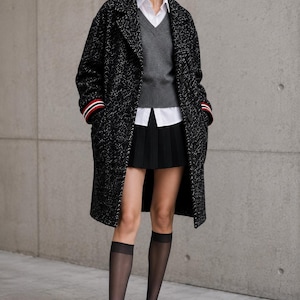 May include: A black and white tweed coat with red, white, and blue striped cuffs, worn over a grey V-neck jumper, a white collared shirt, and a black pleated mini skirt. The outfit includes sheer black knee-high socks and black leather loafers.