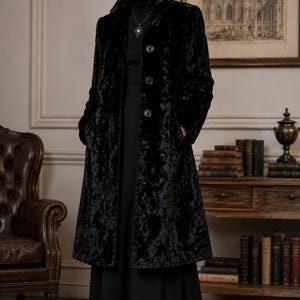 Può includere: Un cappotto nero testurizzato con collo alto, maniche lunghe e chiusura frontale con bottoni. Il cappotto è indossato sopra un top nero a collo alto e una gonna lunga nera, abbinato a stivaletti neri a punta e una collana con pendente.