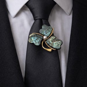 Könnte beinhalten: Nahaufnahme einer schwarzen Krawatte mit einer goldenen und schillernden Brosche. Die Brosche hat die Form eines dreiblättrigen Kleeblatts mit einer kleinen Perle in der Mitte. Die Brosche ist an einer schwarzen Krawatte befestigt, die Person trägt ein weißes Hemd und einen schwarzen Anzug.