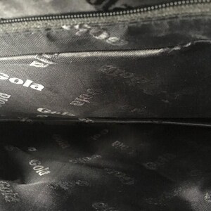 May include: Interior of a black bag with a zipper and a dark gray lining. The lining is patterned with the white word "Gola" repeated throughout. The bag's interior is visible.