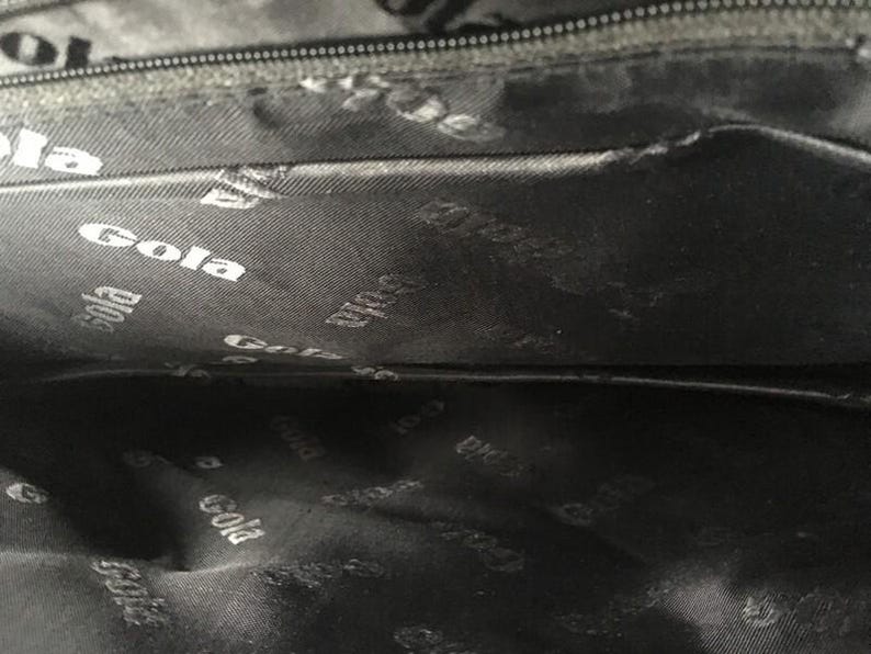 May include: Interior of a black bag with a zipper and a dark gray lining. The lining is patterned with the white word "Gola" repeated throughout. The bag's interior is visible.