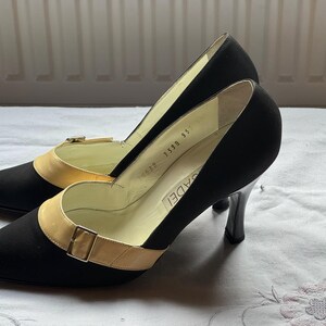 May include: A pair of black high-heeled shoes with a pointed toe and a contrasting tan strap across the vamp, secured with a small buckle. The heels are black and the shoes are set on a white surface.