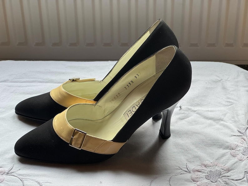 May include: A pair of black high-heeled shoes with a pointed toe and a contrasting tan strap across the vamp, secured with a small buckle. The heels are black and the shoes are set on a white surface.